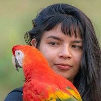 Foto de Txai Suruí, uma jovem indígena de cabelos escuros e lisos. Ela tem franja e segura na foto uma arara.