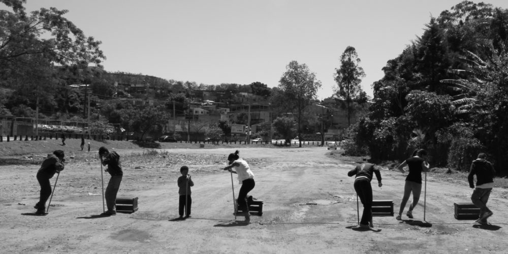 Foto em preto e branco de um grupo de crianças brincando, em um parque de terra, com alguns caixotes e pedaços de madeira. Ao fundo muitas árvores.