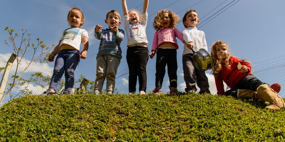 Um grupo de crianças está sobre um monte coberto de grama. Elas sorriem animadas enquanto uma delas até joga as mãos para cima feliz.