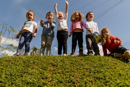 Um grupo de crianças está sobre um monte coberto de grama. Elas sorriem animadas enquanto uma delas até joga as mãos para cima feliz.