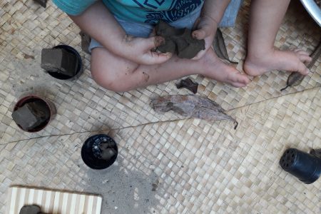 As mãos de uma criança brincam com massa de barro. Ao seu redor há elementos como folhas secas e vasos de plantas.