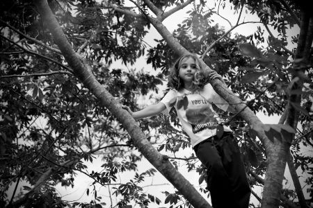 Foto em preto e branco. Criança em cima de uma árvore. Ela está em pé, segurando com as duas mãos em galhos.