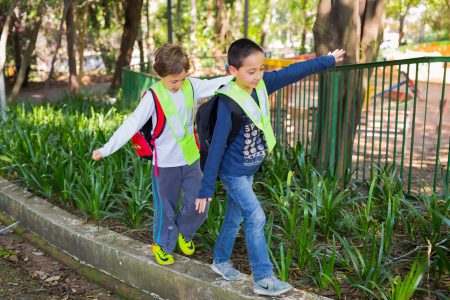 Duas crianças andando sobre uma guia, com os braços abertos para se equilibrar. Ao fundo vegetação e uma grade verde.