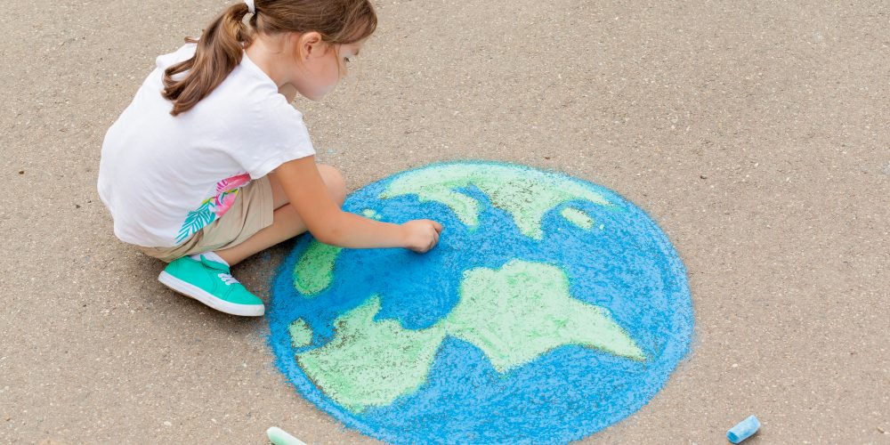 Criança desenha o planeta terra no chão, usando giz de lousa nas cores azul e verde.