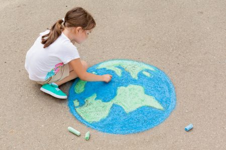 Criança desenha o planeta terra no chão, usando giz de lousa nas cores azul e verde.
