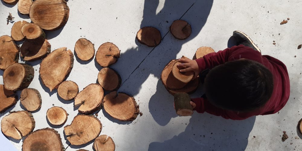 Criança, vista de cima, brinca com pedaços de madeira cortados em forma circular.