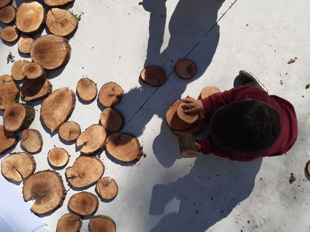 Criança, vista de cima, brinca com pedaços de madeira cortados em forma circular.