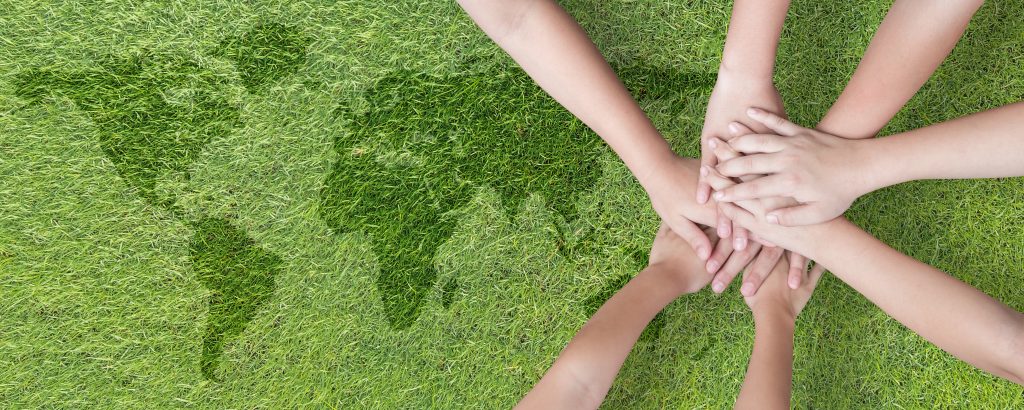 Foto tirada do ângulo de cima das mãos de crianças unidas. Embaixo e ao lado das mãos é possível ver o mapa mundi em um gramado.