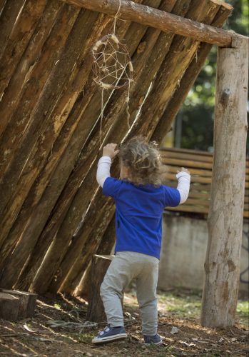 Criança pequena brincando com um filtro do sonho pendurado em uma cabana feita de madeira.