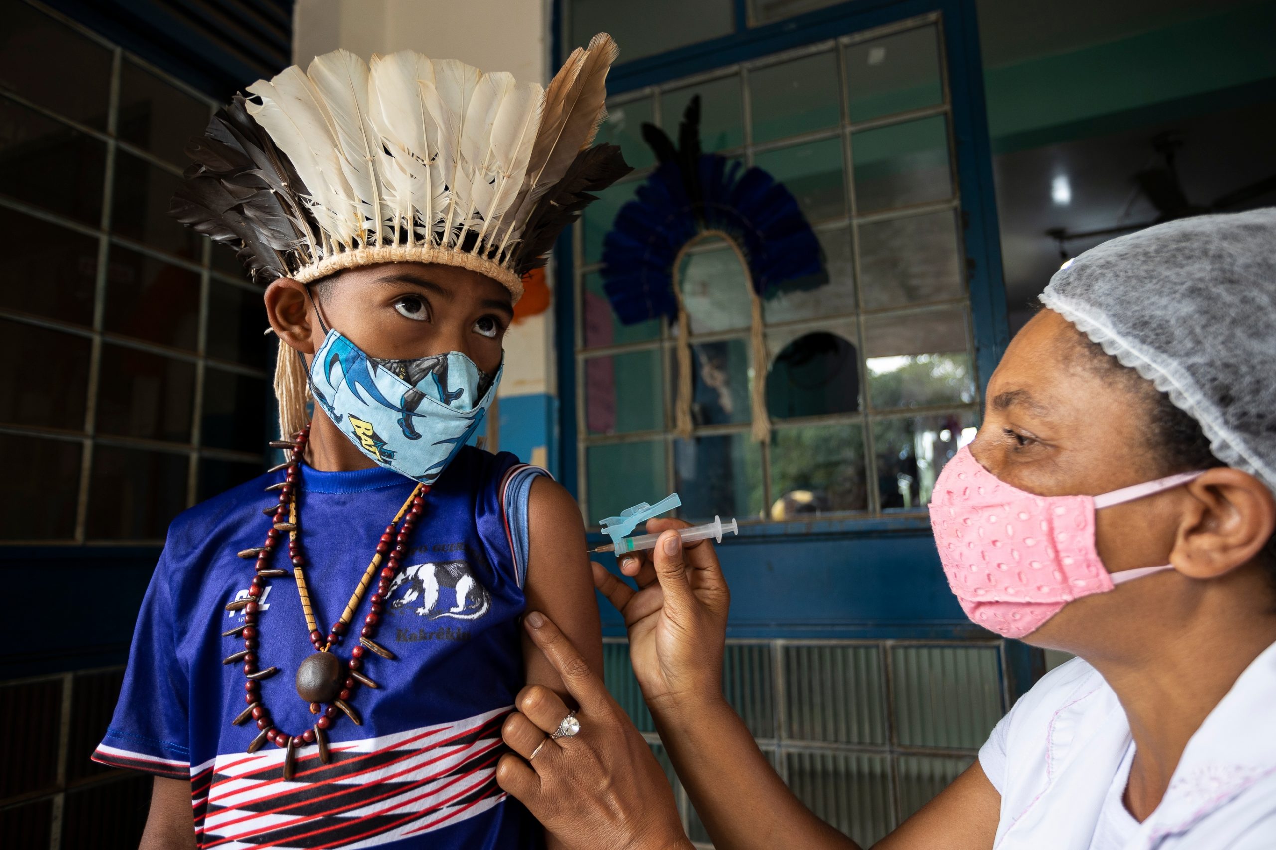Criança indígena, usando mascará e adereços de sua tribo, sendo vacinada por uma enfermeira.