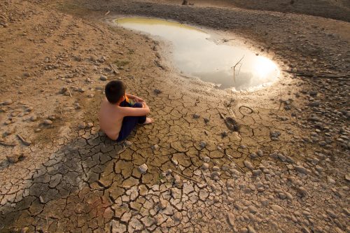 Criança sentada sobre solo seco e rachado, observando o que restou de água. 