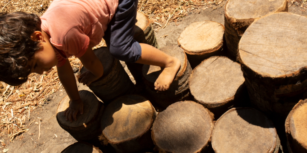 Criança brincando sobre troncos de madeira de diferentes tamanhos e agrupados.