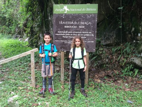 Crianças com mochilas e equipamentos para fazer trilhas em frente a placa do Parque Nacional do Itatiaia