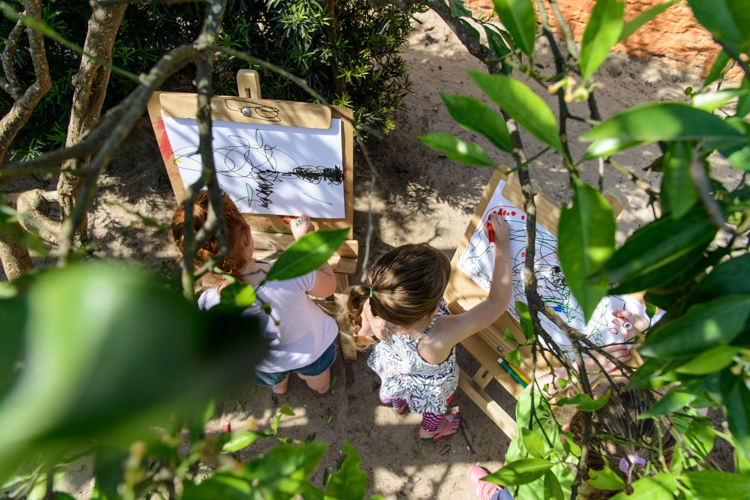 Imagem vista de cima mostra duas crianças pintando em cavalete, ao ar livre e na sombra.