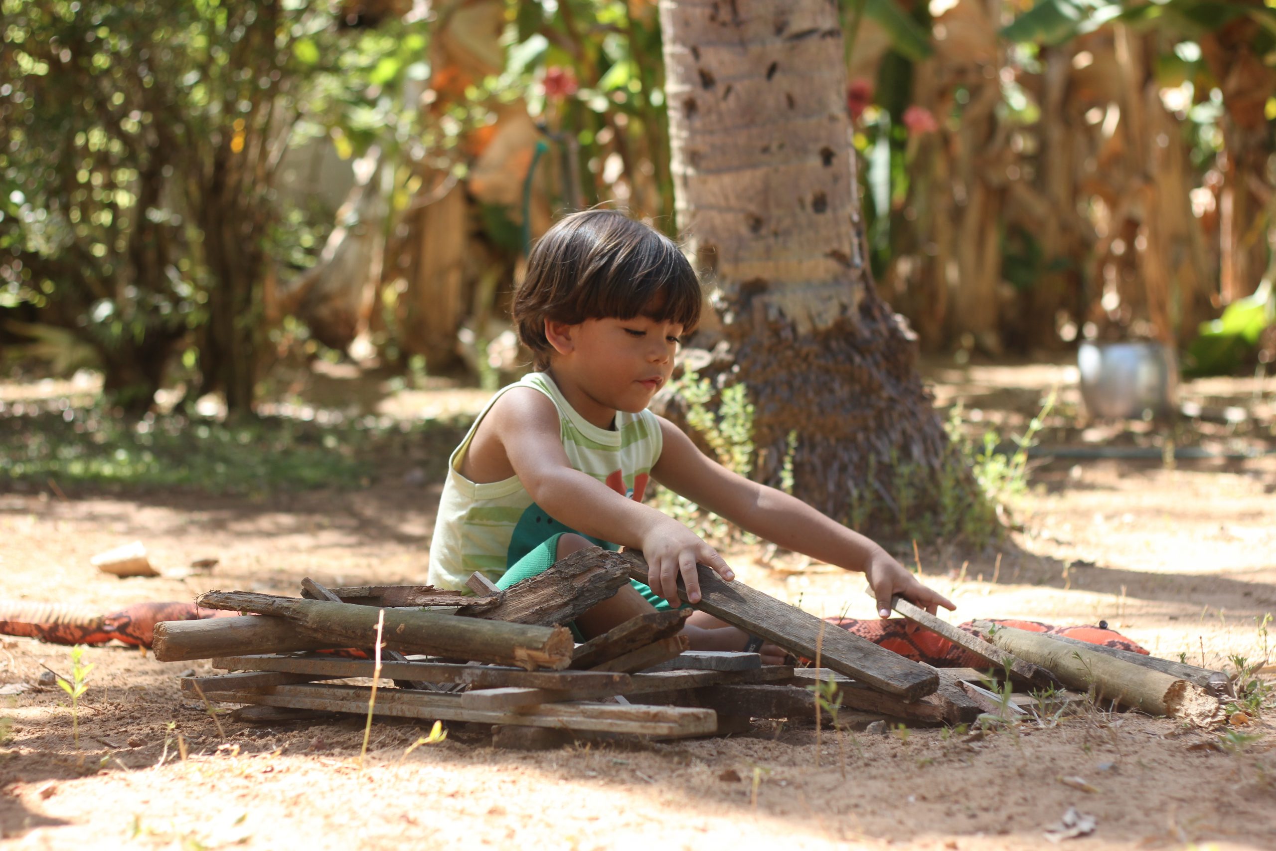 Criança brincando com madeiras, sentada no chão de terra e ao ar livre. Manifesto por escolas com mais natureza
