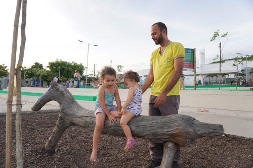 Duas meninas, acompanhadas do pai, brincam sentadas em uma escultura de madeira que lembra um cavalo. Parque naturalizado em Caruaru.