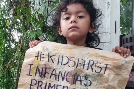 Criança segura uma folha de papel, onde está escrito “#KidsFirst, Crianças primeiro