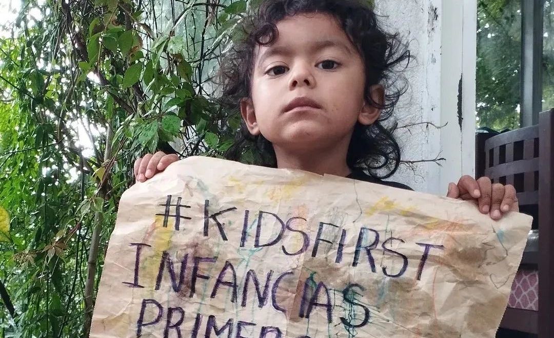 Criança segura uma folha de papel, onde está escrito “#KidsFirst, Crianças primeiro