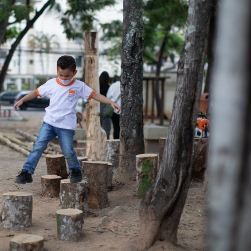 Foto mostra um menino usando jeans e tênis, com camiseta e máscara facial da rede pública de ensino de Niterói, Rio de Janeiro, caminhando sobre tocos de árvores fincados no chão de terra, em uma ampla área livre, arborizada. Ao fundo algumas pessoas, carros e construções.