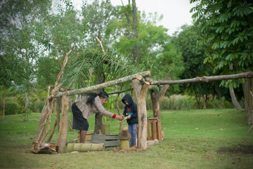 Foto mostra uma ampla área verde gramada e arborizada. Ao centro, uma cabana feita com diversos troncos fincados no chão, com o telhado feito de folhas de palmeira. Dentro da cabana uma senhora e um menino brincam com objetos naturais.