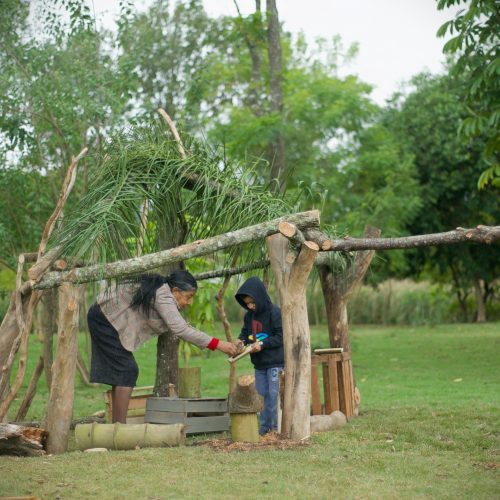Foto mostra uma ampla área verde gramada e arborizada. Ao centro, uma cabana feita com diversos troncos fincados no chão, com o telhado feito de folhas de palmeira. Dentro da cabana uma senhora e um menino brincam com objetos naturais.