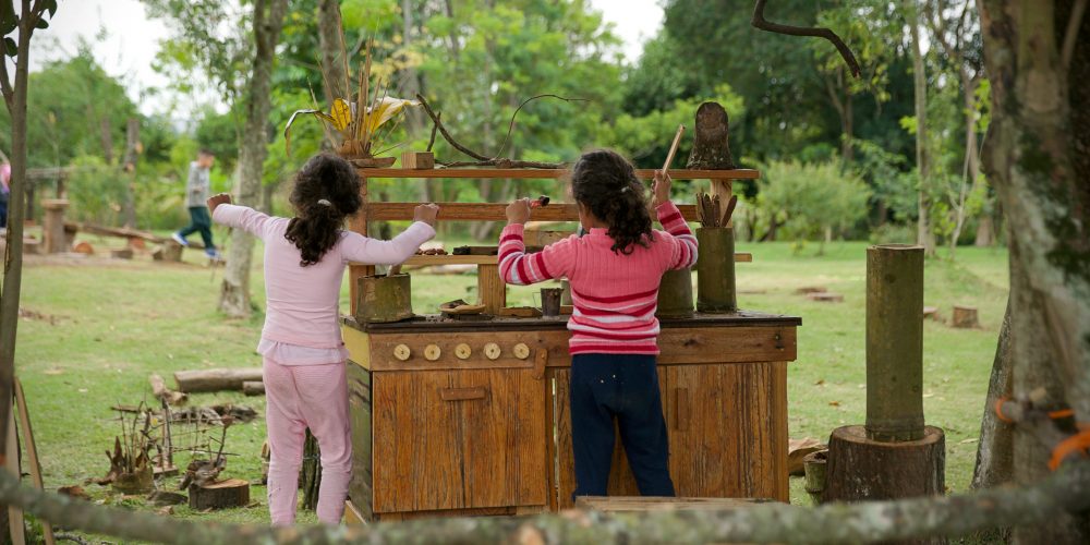 Foto mostra duas meninas, de costas, brincando em uma cozinha da floresta com seus utensílios naturais, instalada numa área verde ampla e arborizada, na qual há outros brinquedos naturalizados. Ao fundo, um menino caminha sobre uma trave de equilíbrio.
