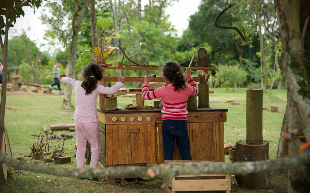 [:pt]Foto mostra duas meninas, de costas, brincando em uma cozinha da floresta com seus utensílios naturais, instalada numa área verde ampla e arborizada, na qual há outros brinquedos naturalizados. Ao fundo, um menino caminha sobre uma trave de equilíbrio.[:]
