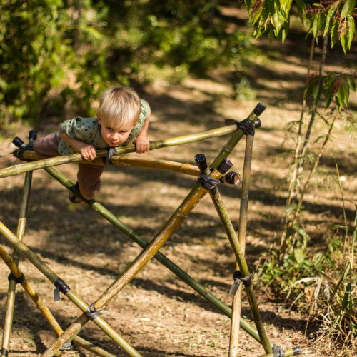 Foto mostra um menino pequeno equilibrando seu corpo no topo de uma estrela de bambu, em uma área verde muito arborizada.