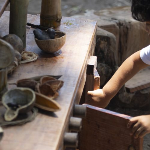 Foto em ângulo fechado de um menino brincando em uma cozinha da floresta. Sobre o móvel, diversos utensílios naturais como bambus, sementes e cascas de coco seco. O menino está abrindo as portas do armário que integra o móvel.