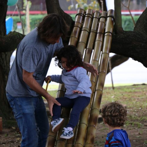 Foto mostra um escorregador feito com cinco hastes de bambu dispostas lado a lado e apoiadas na parte superior por um tronco de árvore. Uma menina pequena escorrega, assistida por um adulto, enquanto outra criança pequena, vista de costas, observa.