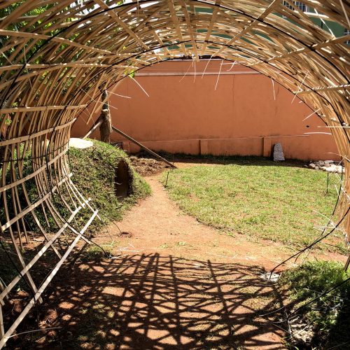 Foto mostra a vista interna da estrutura de tiras de bambu de um túnel vivo, montada num quintal de chão de terra e grama. Ao fundo, se vê um muro terracota.