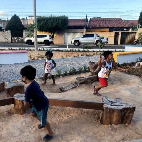 Foto mostra três crianças correndo ao redor de um brinquedo de madeira feito com traves, troncos e tocos de árvores instalados sobre um chão de areia. Ao fundo, um canteiro, uma rua com muitas casas e dois carros estacionados.