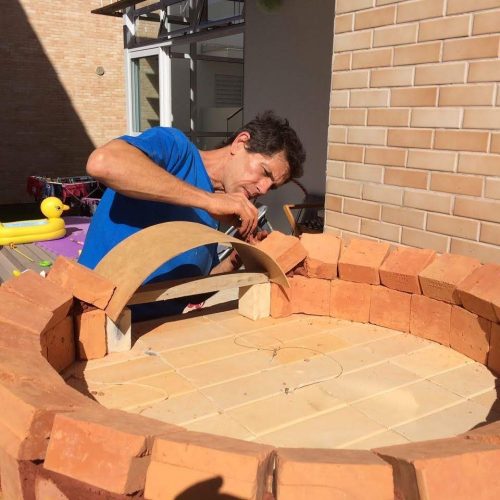 Foto mostra um forninho de barro em construção, com um homem adulto encaixando um tijolo em sua base.