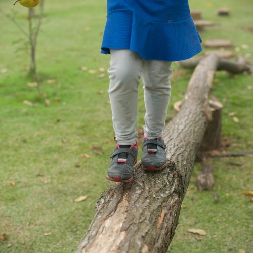 Foto de uma área gramada onde uma criança, vista da cintura para baixo, usando vestido azul, legging e tênis, caminha sobre um trepa-tronco horizontal que está apoiado no chão e em tocos de madeira