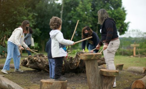 Crianças e adultos brincando com baquetas em uma brinquedo natural, feito com podas de árvores.