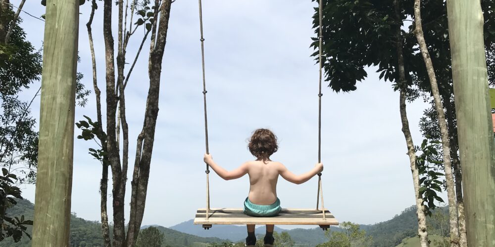 De costas, uma criança se balança entre duas árvores, olhando para um ambiente natural