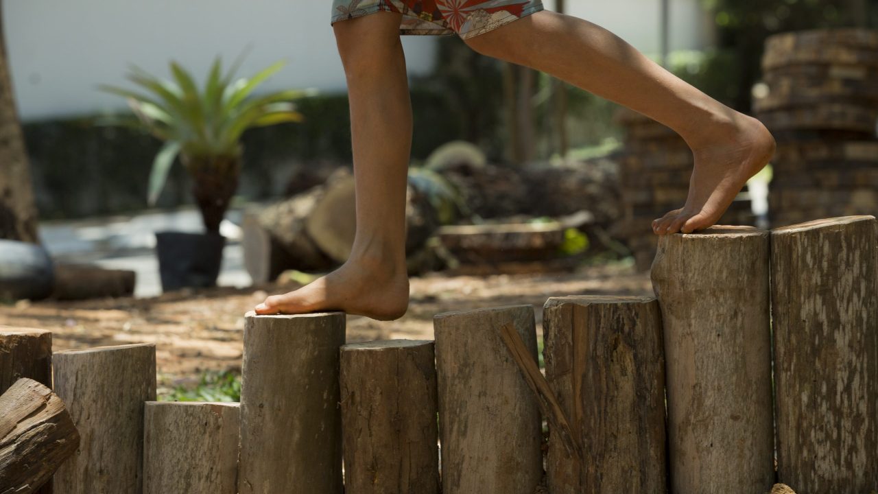Pés descalços de uma criança caminham sobre troncos de madeira cortados em tamanhos diferentes.